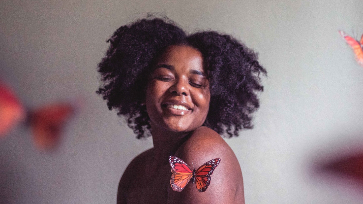 Woman with afro hair smiling surrounded by butterflies on a grey background. 