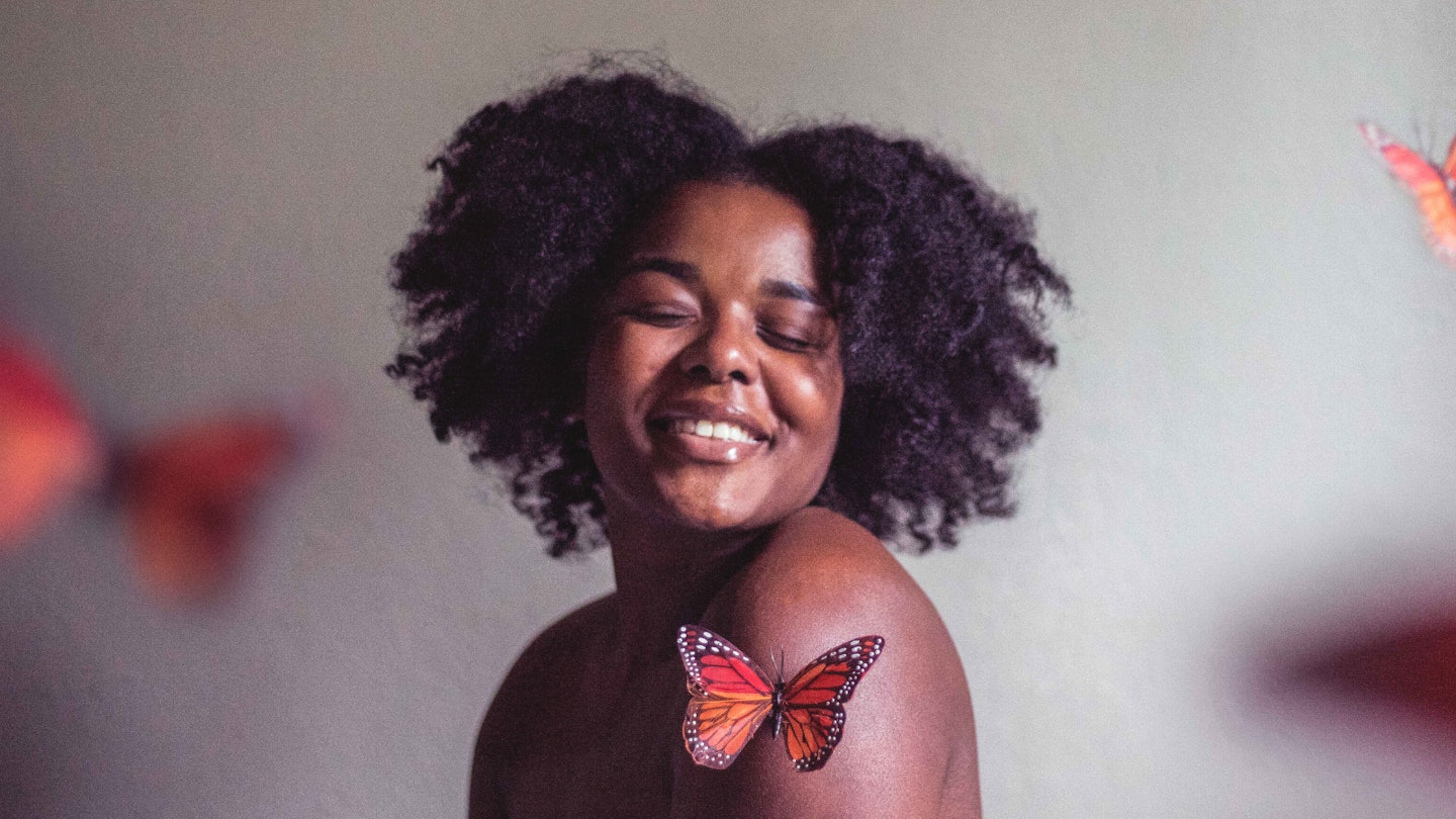 Woman with afro hair smiling surrounded by butterflies on a grey background.