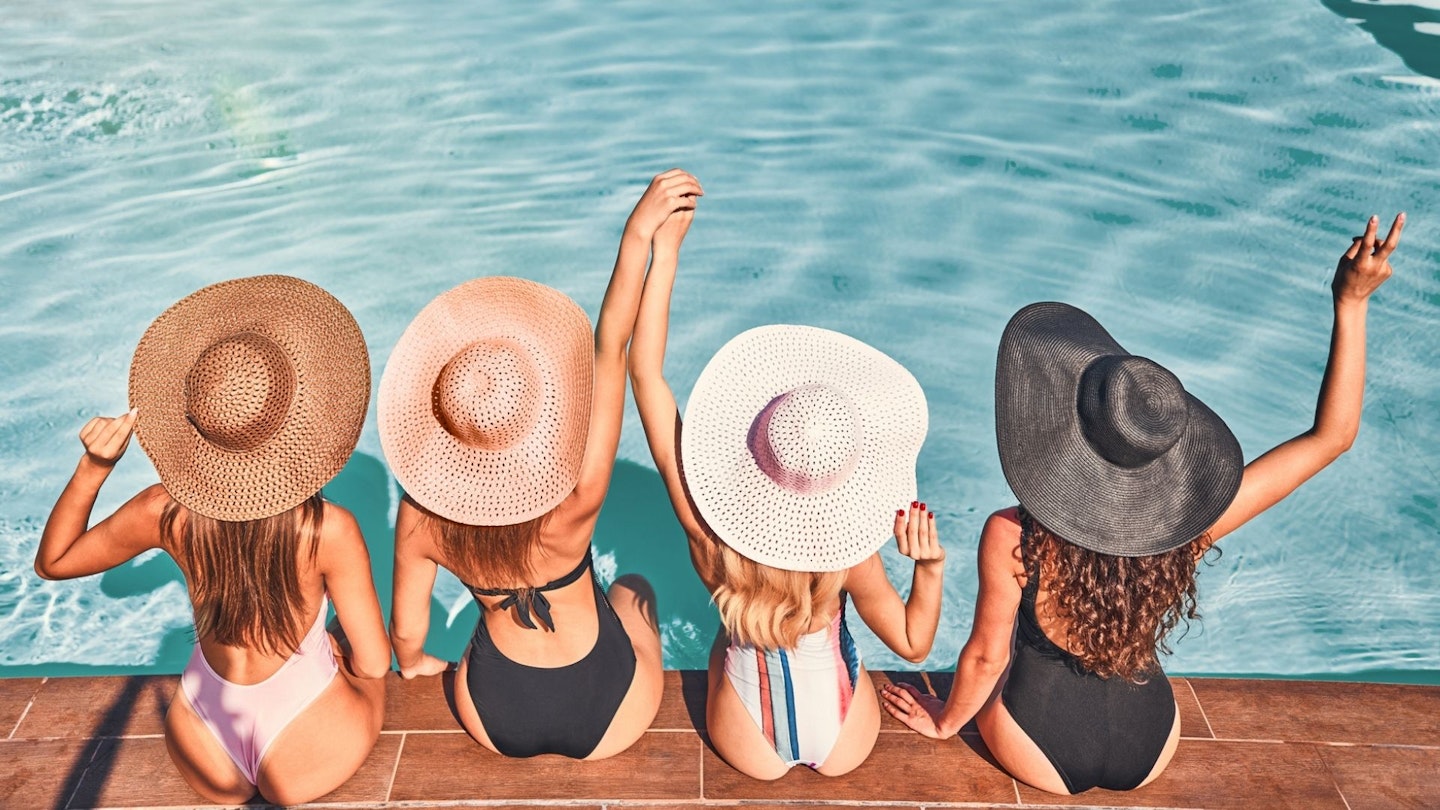 Holiday checklist: Four young women with sun hats sitting by the poolside of a resort swimming pool during a summer holiday
