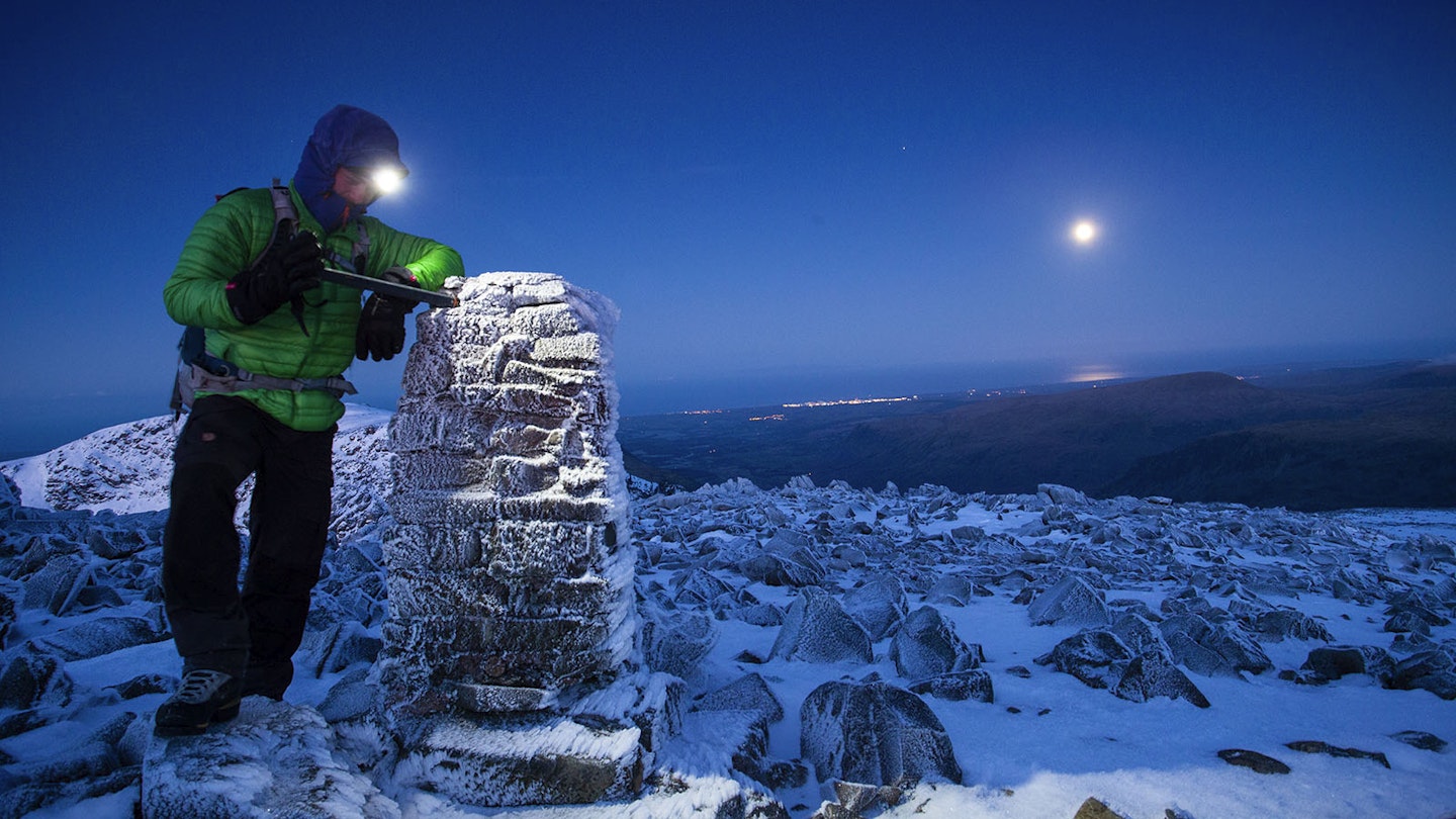 Man with head torch and ice axe on winter mountain with trig point