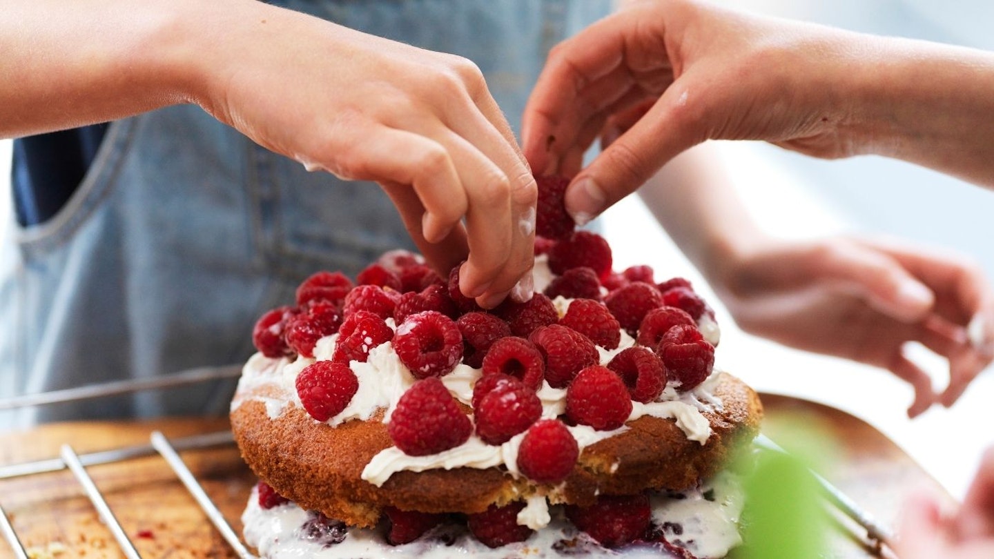 Girl and her sister baking a cake, decorating cake with fresh cream and raspberries at kitchen table, cropped view of hands.