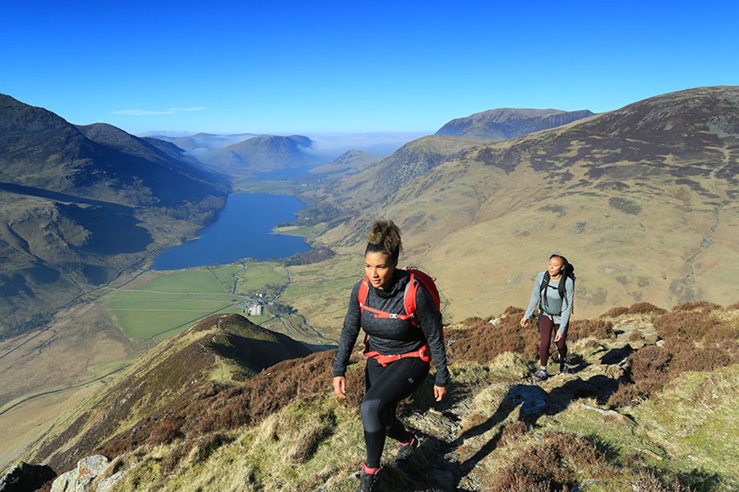 Two women in walking and hiking gear ascending Fleetwith Pike in the Lake District with Buttermere behind them