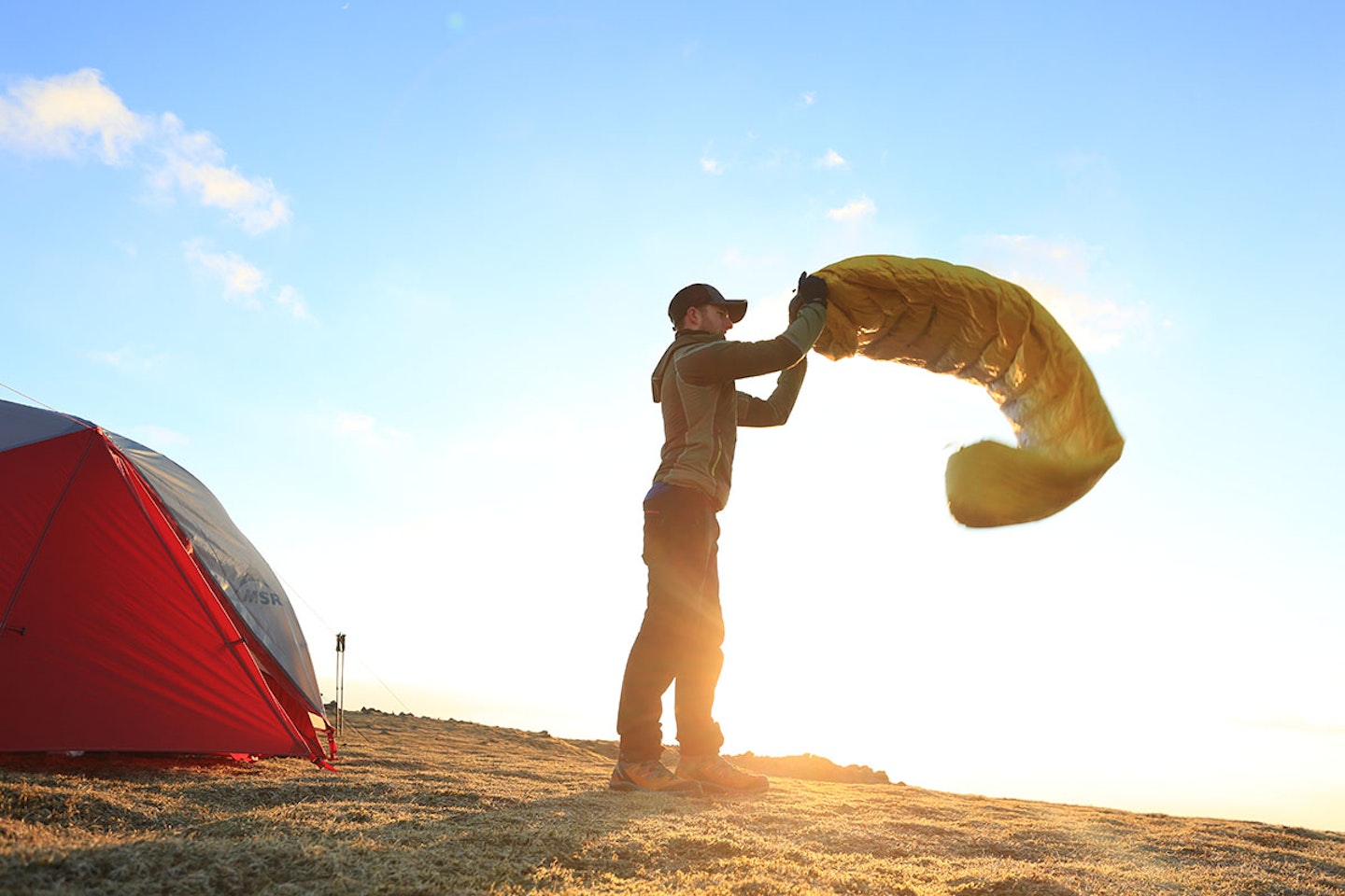 Man shaking out sleeping bag with tent and sky in the mountains