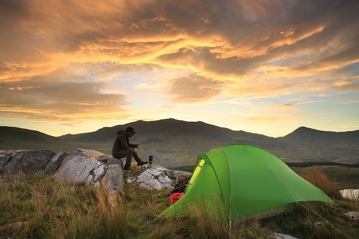 man on rock camping with stove and tent at sunset in the mountains
