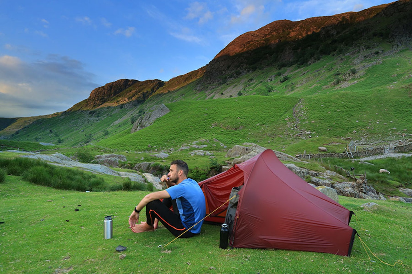 man brushing his teeth outside tent camping on grass in a valley with cliffs behind