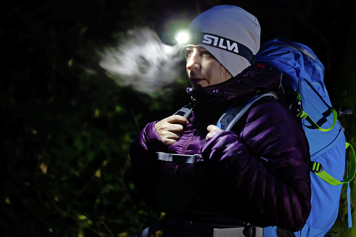 woman in hiking gear and hat and rucksack in dark with head torch illuminating her breath