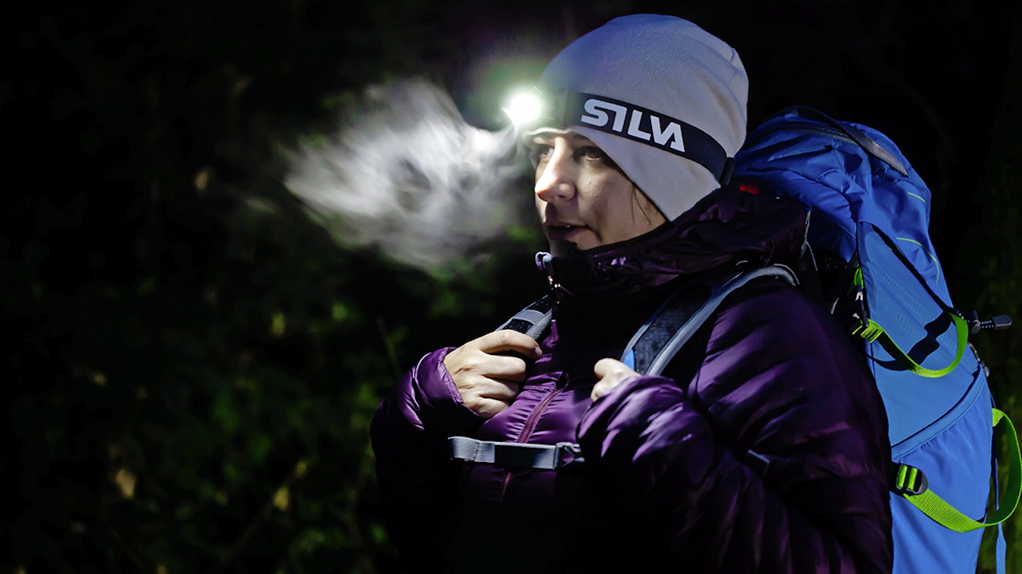 woman in hiking gear and hat and rucksack in dark with head torch illuminating her breath