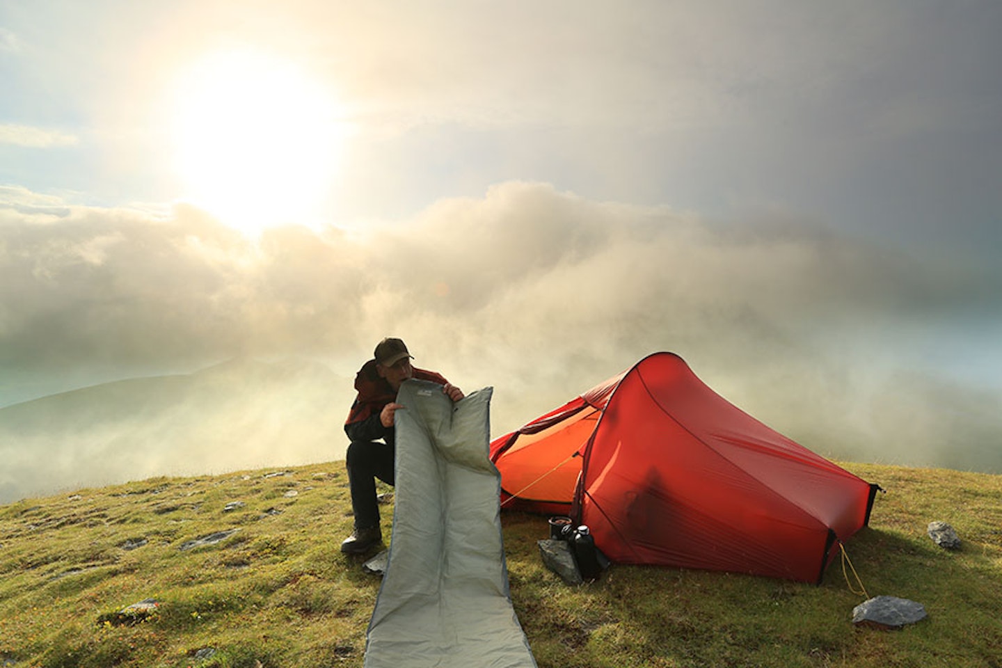 man inflating a sleeping mat by a tent on a hill above a cloud inversion