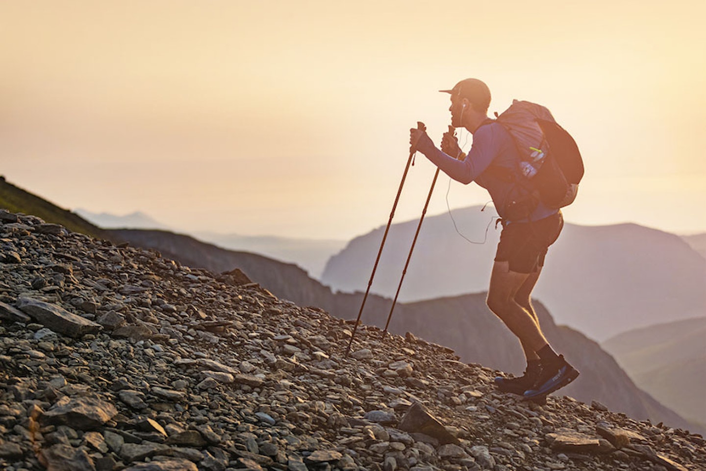 Man walking up a mountain in shorts and cap with headphones rucksack and trekking poles at sunset or sunrise