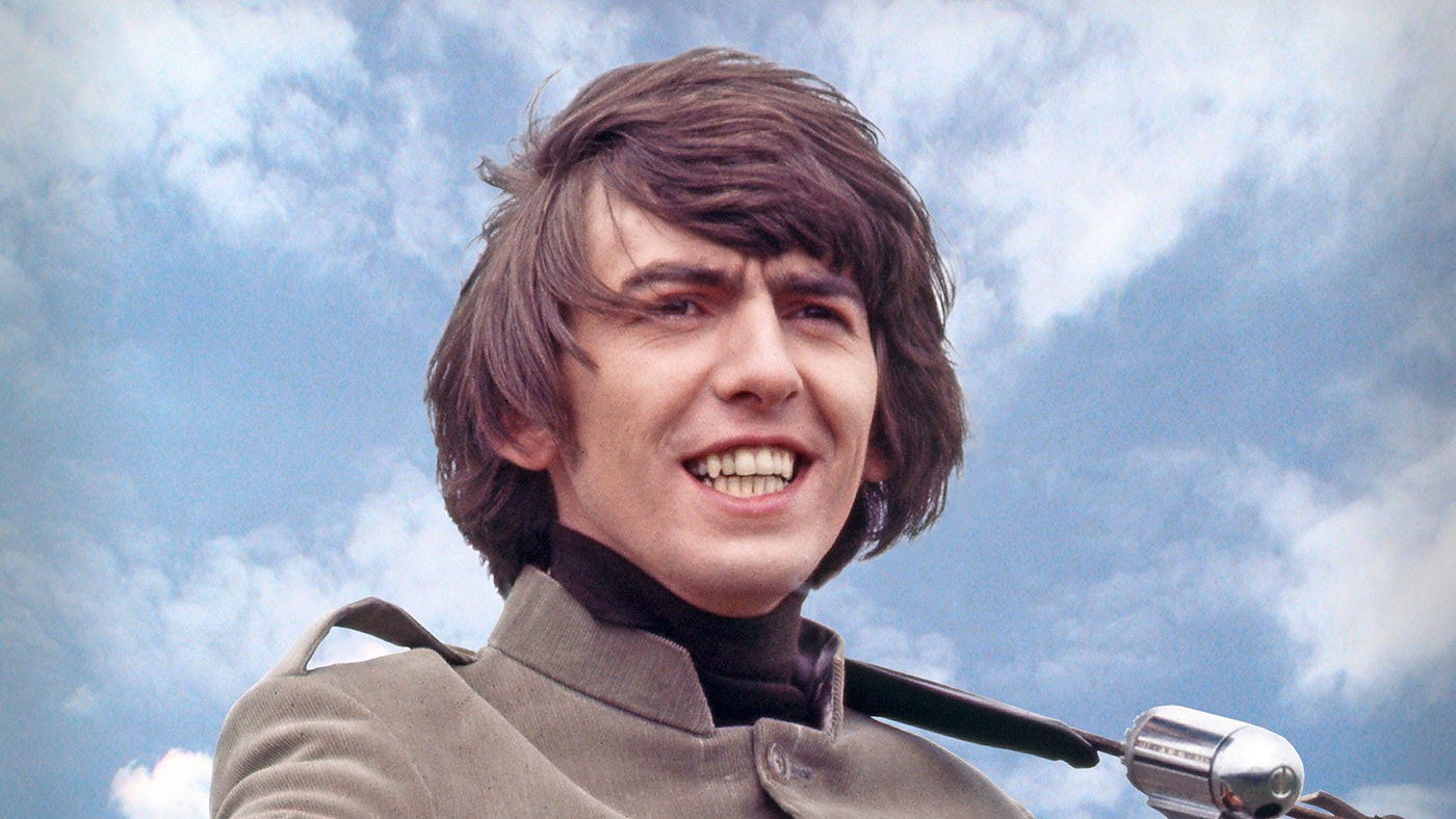 Young George Harrison of The Beatles smiling in a grey jacket with a black turtleneck, standing against a blue sky with light clouds.