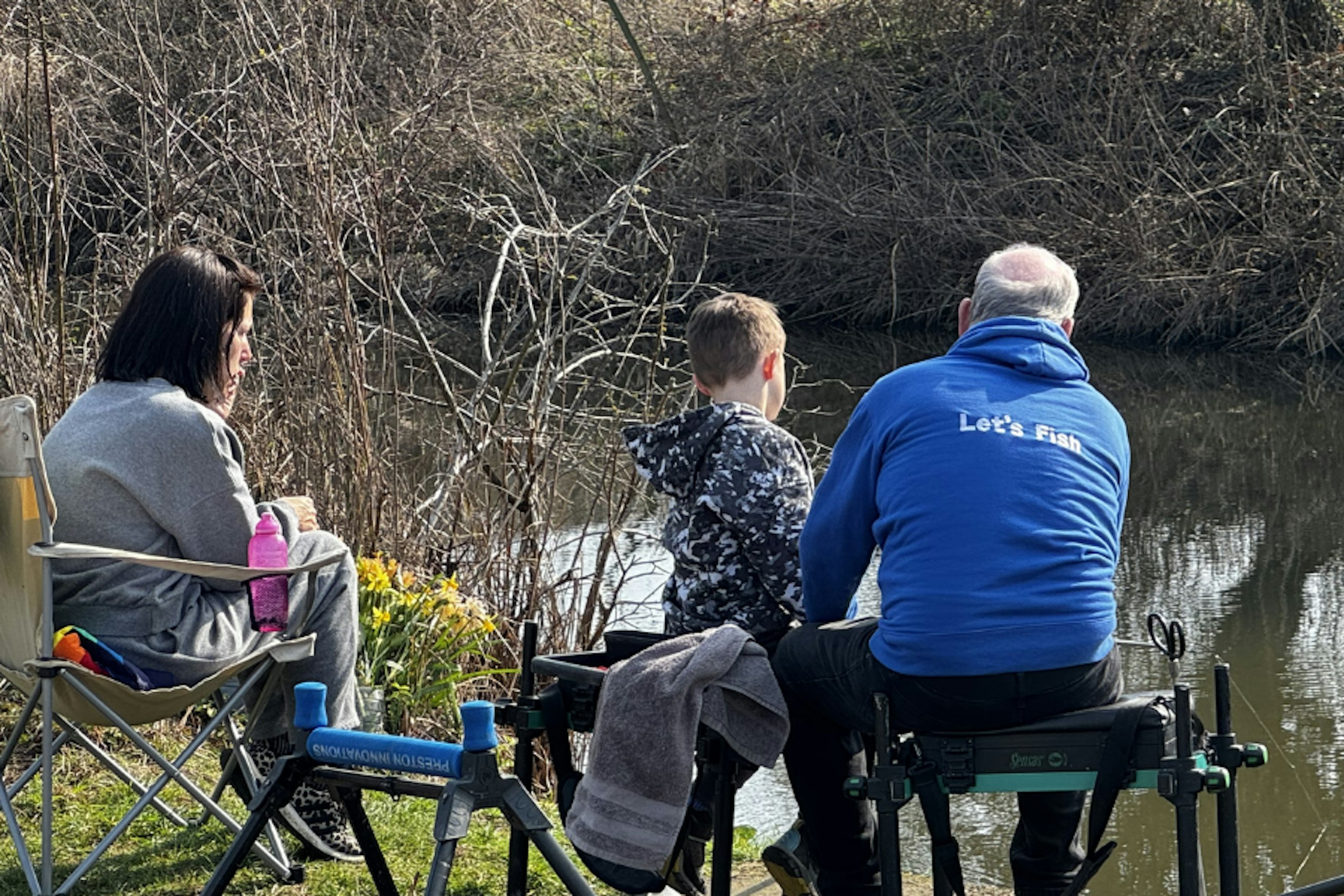 A parent watches as a child fishes alongside a Let’s Fish coach on a small canal, viewed from behind.
