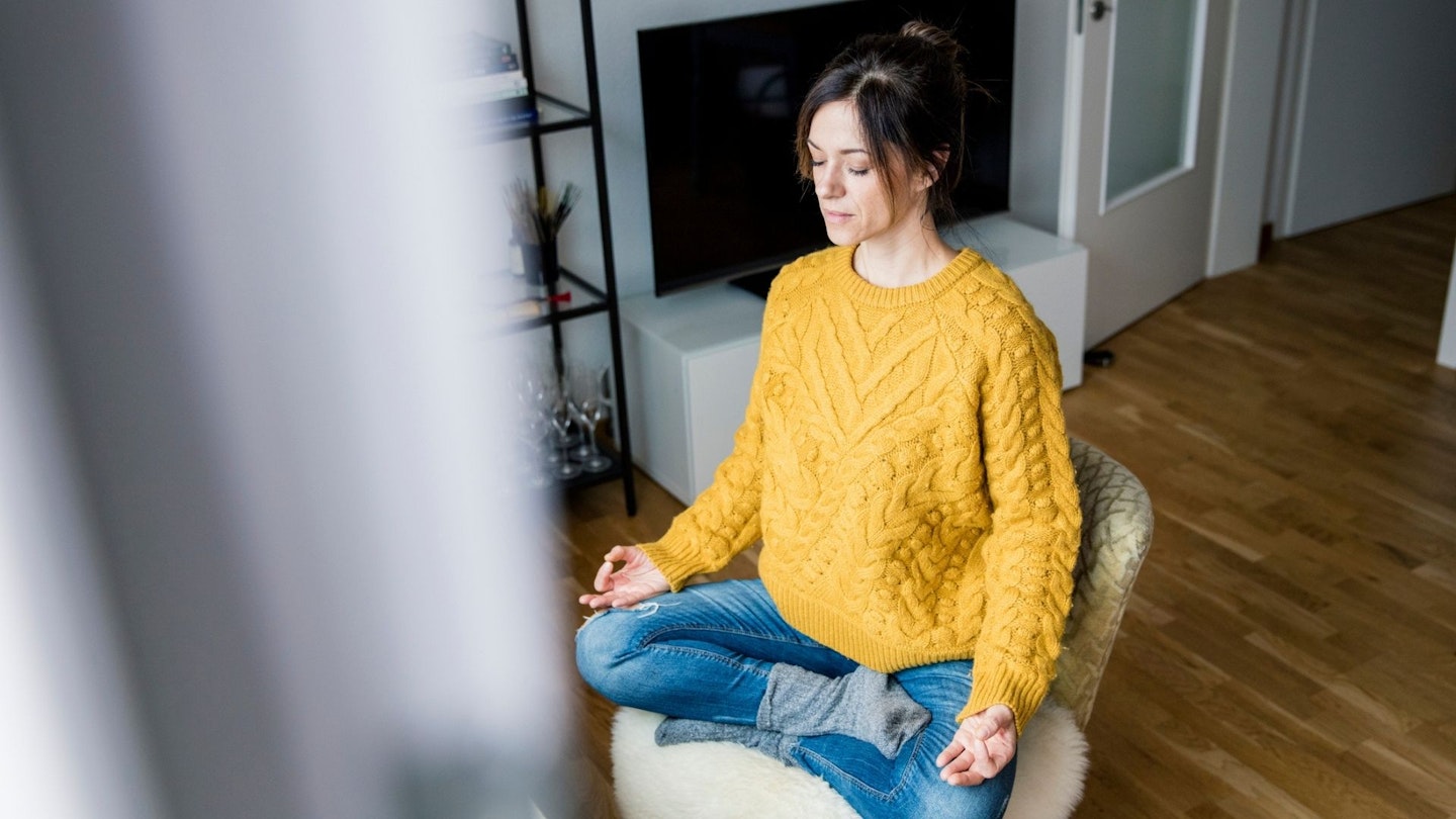 Mature woman sitting cross-legged on chair with eyes closed, meditating at home